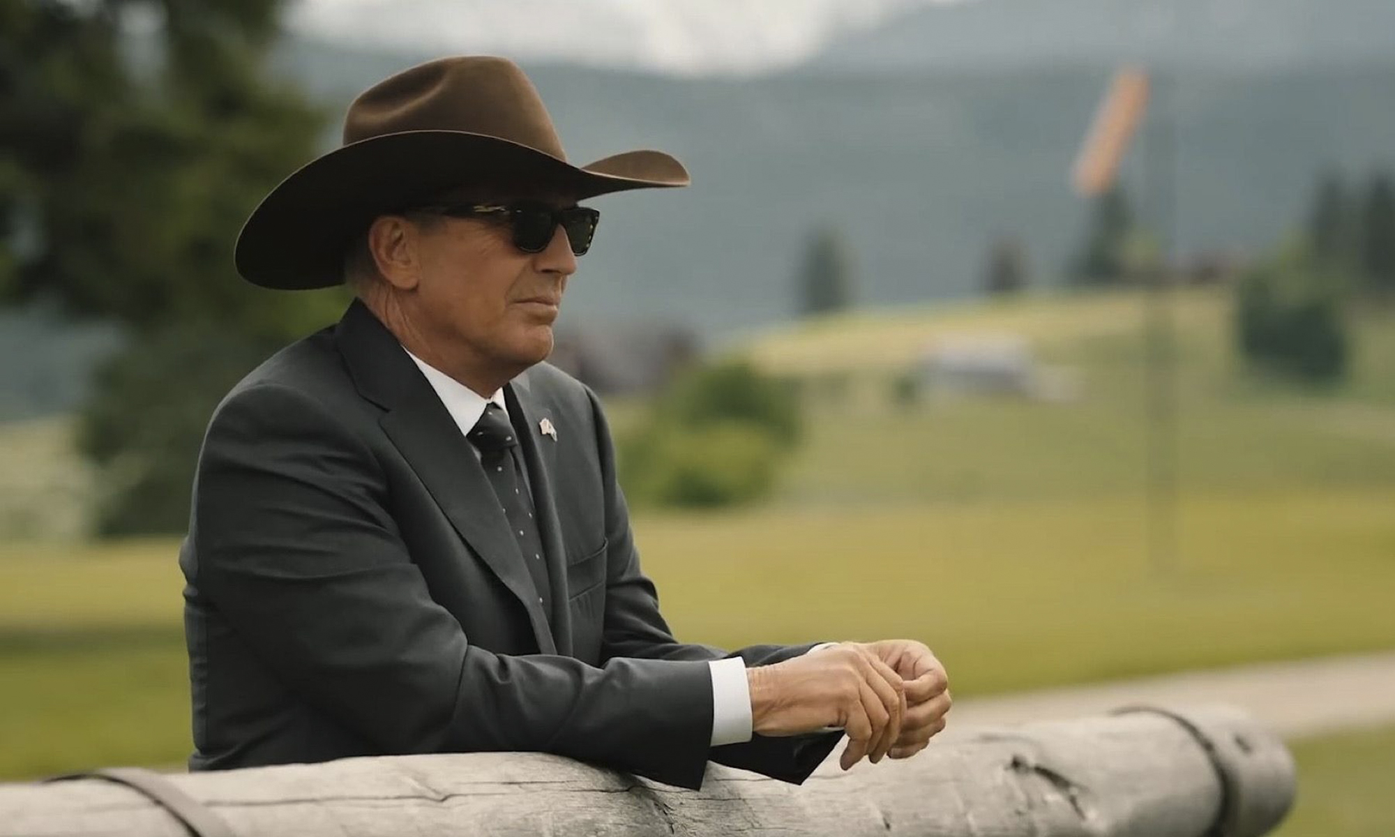 John Dutton de Yellowstone descansando sobre una valla y mirando hacia afuera con un sombrero de vaquero y gafas de sol.