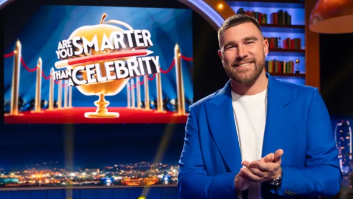 Travis Kelce está com as mãos cruzadas em um cenário de game show.