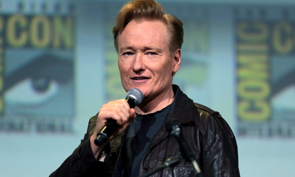 Conan O'Brien holds a mic up by his mouth.