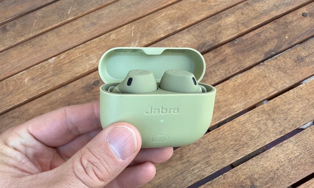 A hand holding olive colored Jabra Elite 8 Active earbuds in their open case above a wooden outdoor table.