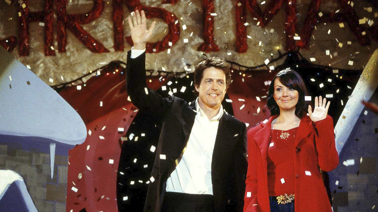 A man and woman stand on stage waving as confetti falls around them in Love Actually. 