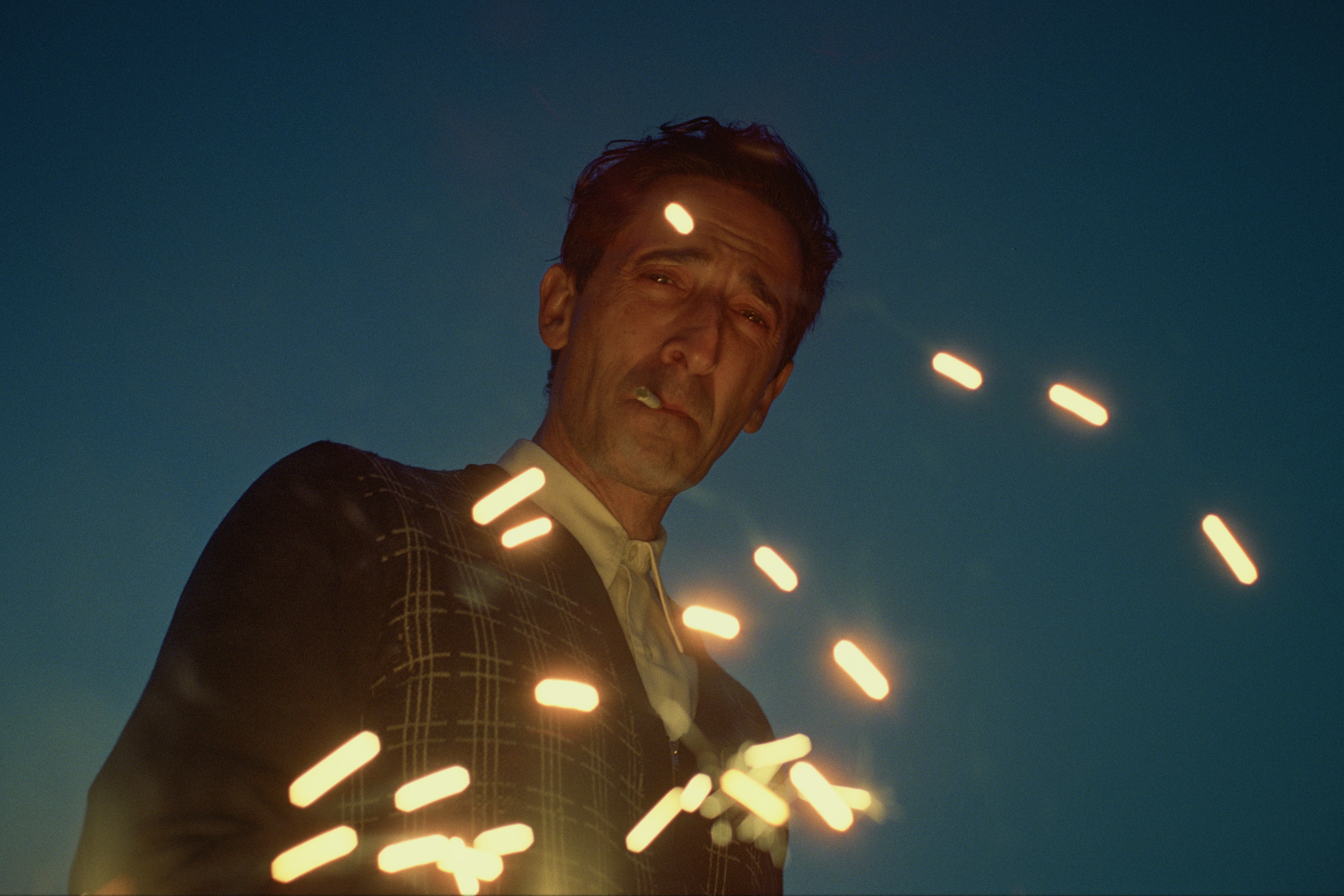 Adrien Brody smokes as sparks dance around him in a still from the movie The Brutalist