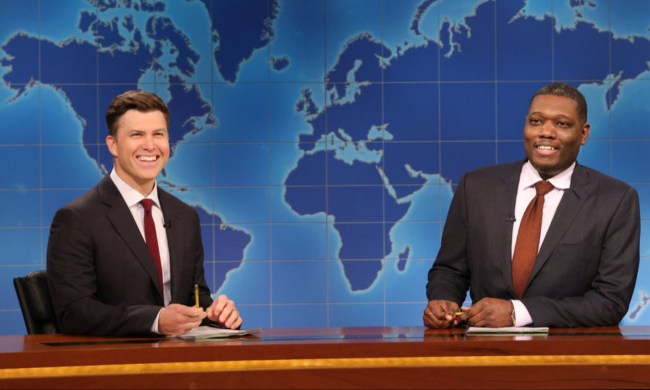 Colin Jost and Michael Che sit at a desk and laugh on Saturday Night Live.