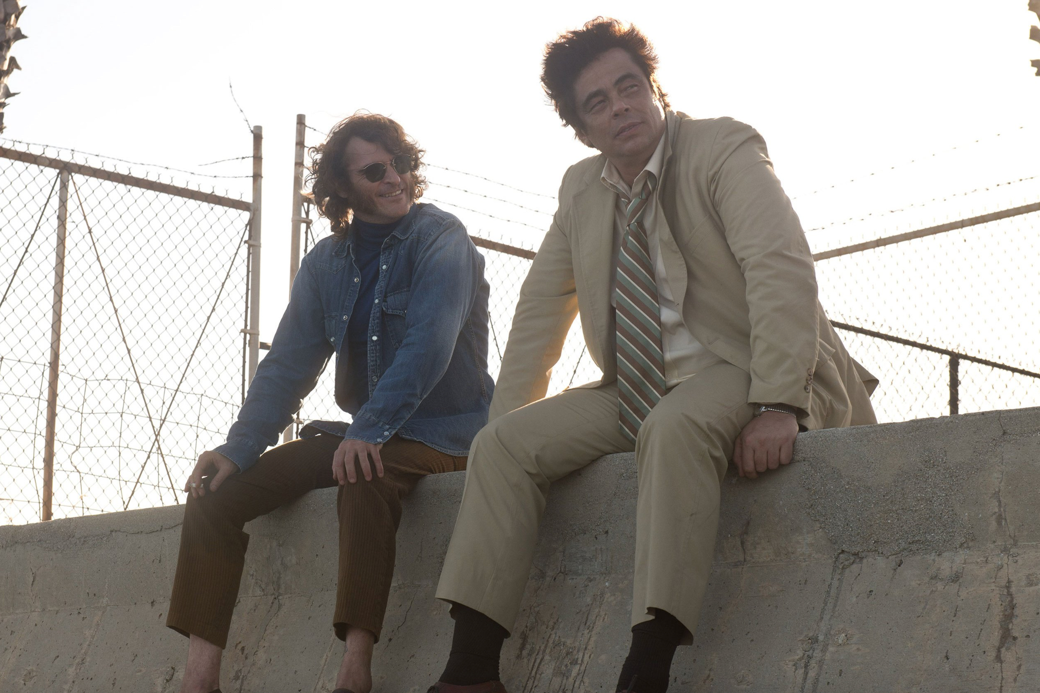 Joaquin Phoenix e Benicio Del Toro sentam-se em uma divisória de concreto em Inherent Vice.