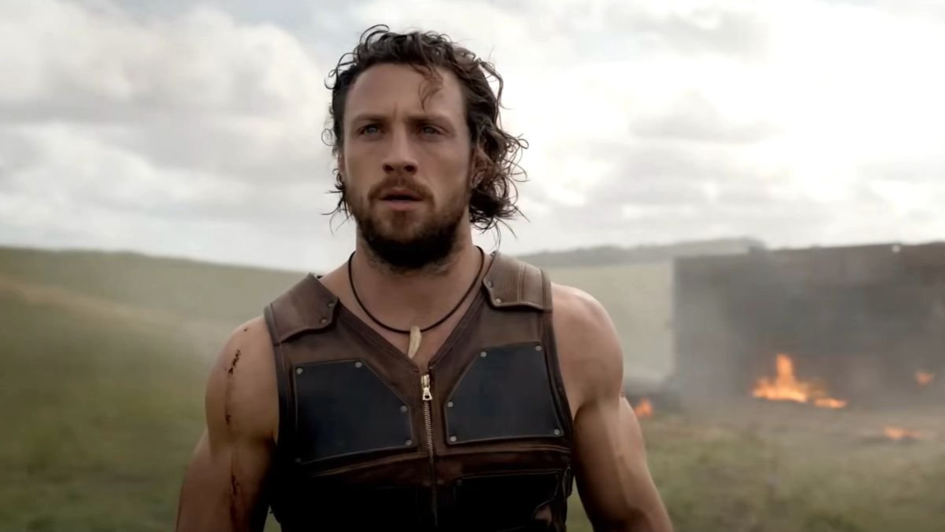 Aaron Taylor-Johnson looks sternly forward as a small fire rages behind him in a still from the movie Kraven the Hunter.