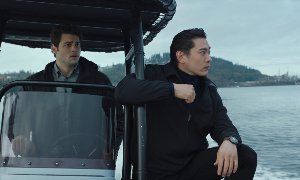 A man drives a boat with another man standing in front of him and staring.