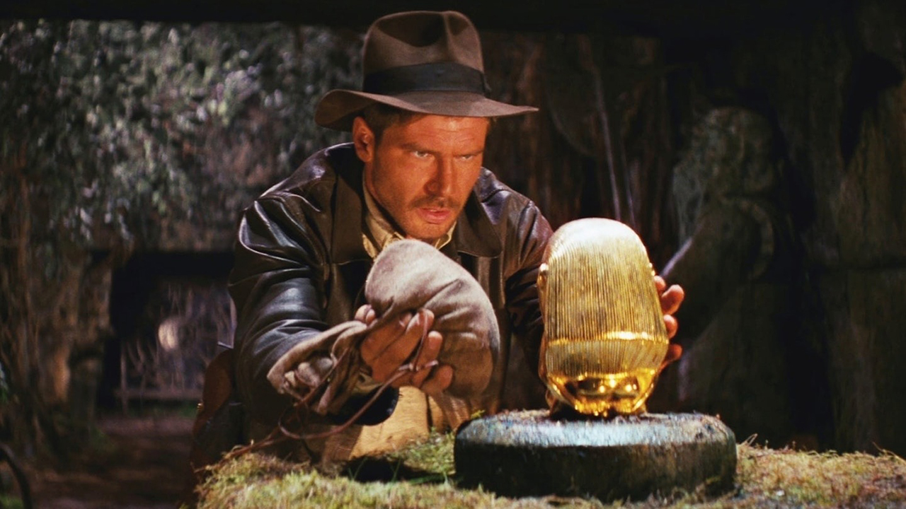 Indiana Jones blickt in Raiders of the Lost Ark aufmerksam auf eine Statue, während er eine Tasche in einer Hand hält.