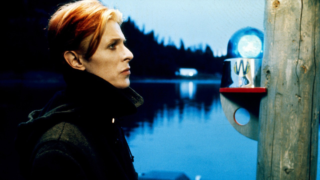 Un profil latéral de David Bowie dans une veste noire, debout au bord de l'eau dans The Man Who Fell To Earth.