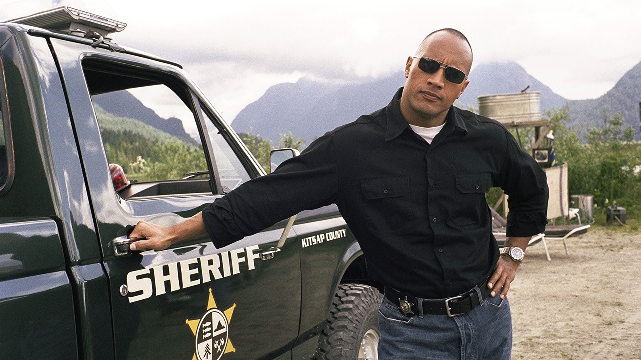 Dwayne Johnson con gafas de sol junto al vehículo del sheriff en Walking Tall