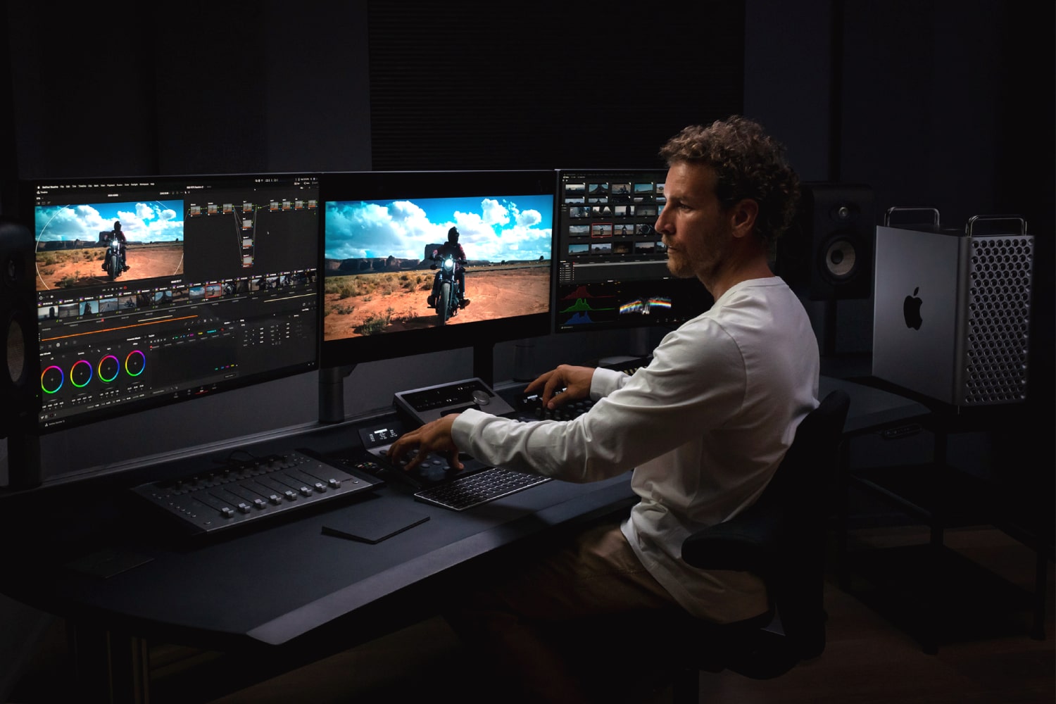 A person uses an Apple Mac Pro along with three monitors and an editing console in a dark room.