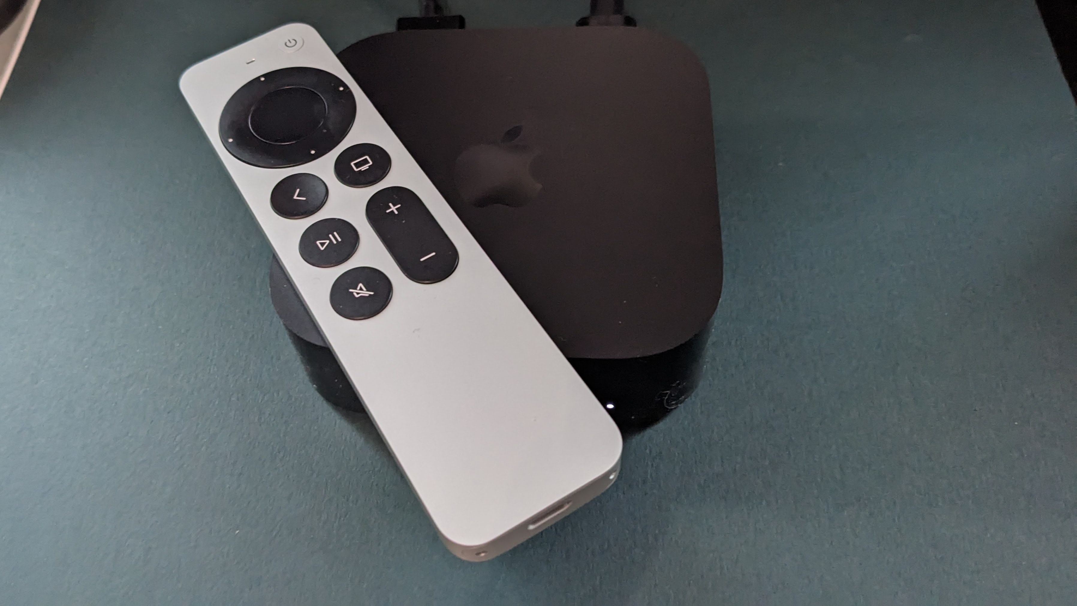 Um Apple TV 4K contra um fundo verde. O controle remoto prateado da maçã fica em cima da caixa de streaming.