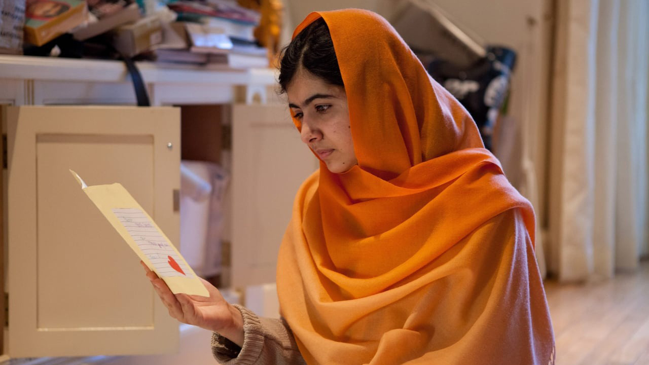 Malala sitting on the floor looking at a paper in He Named Me Malala.