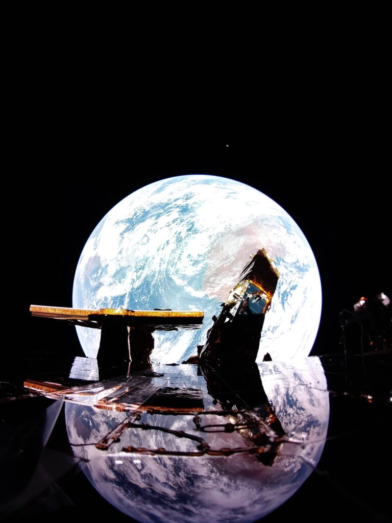 L'atterrisseur Blue Ghost de Firefly capture une image de la Terre se reflétant sur le panneau solaire avec la Lune à l'horizon au-dessus de la Terre. L’antenne en bande X de Firefly et la charge utile LEXI de la NASA sont également présentées sur le pont supérieur de l’atterrisseur.