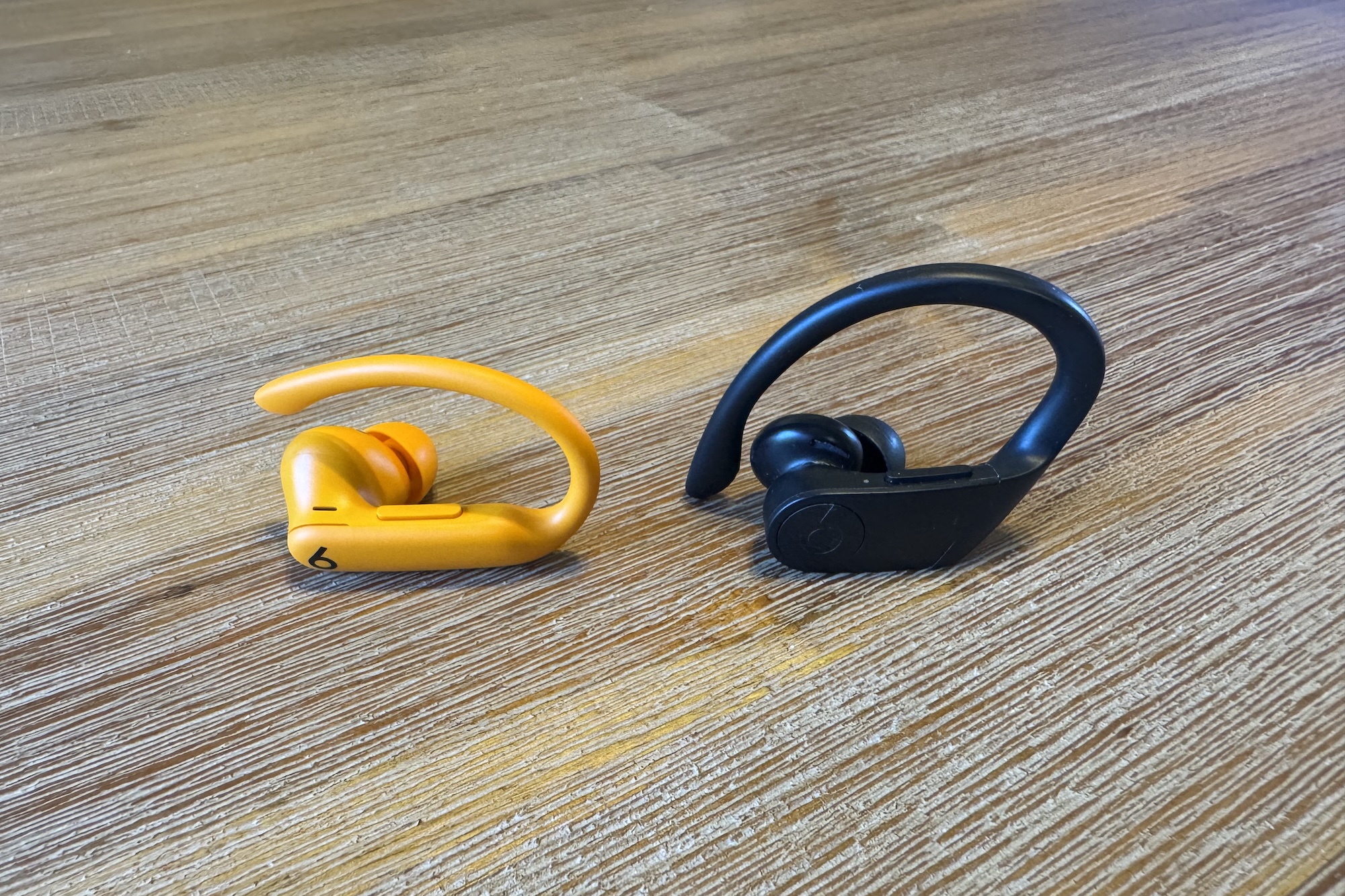 Um fone de ouvido Beats Powerbeats Pro 2 próximo a um fone de ouvido Powerbeats Pro em um piso de madeira.