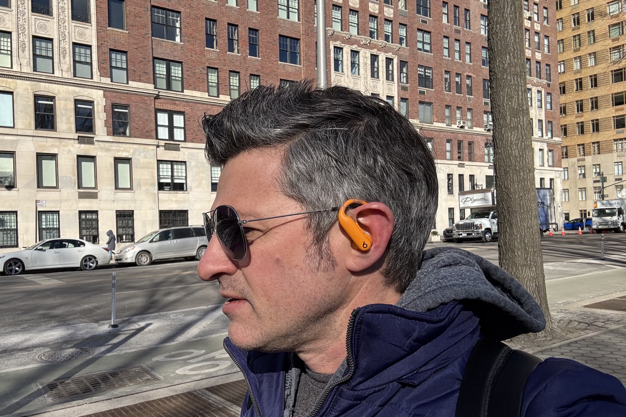 Um homem com uma jaqueta azul e óculos escuros usando fones de ouvido Beats Powerbeats Pro 2 laranja enquanto caminhava por uma rua de Nova York.