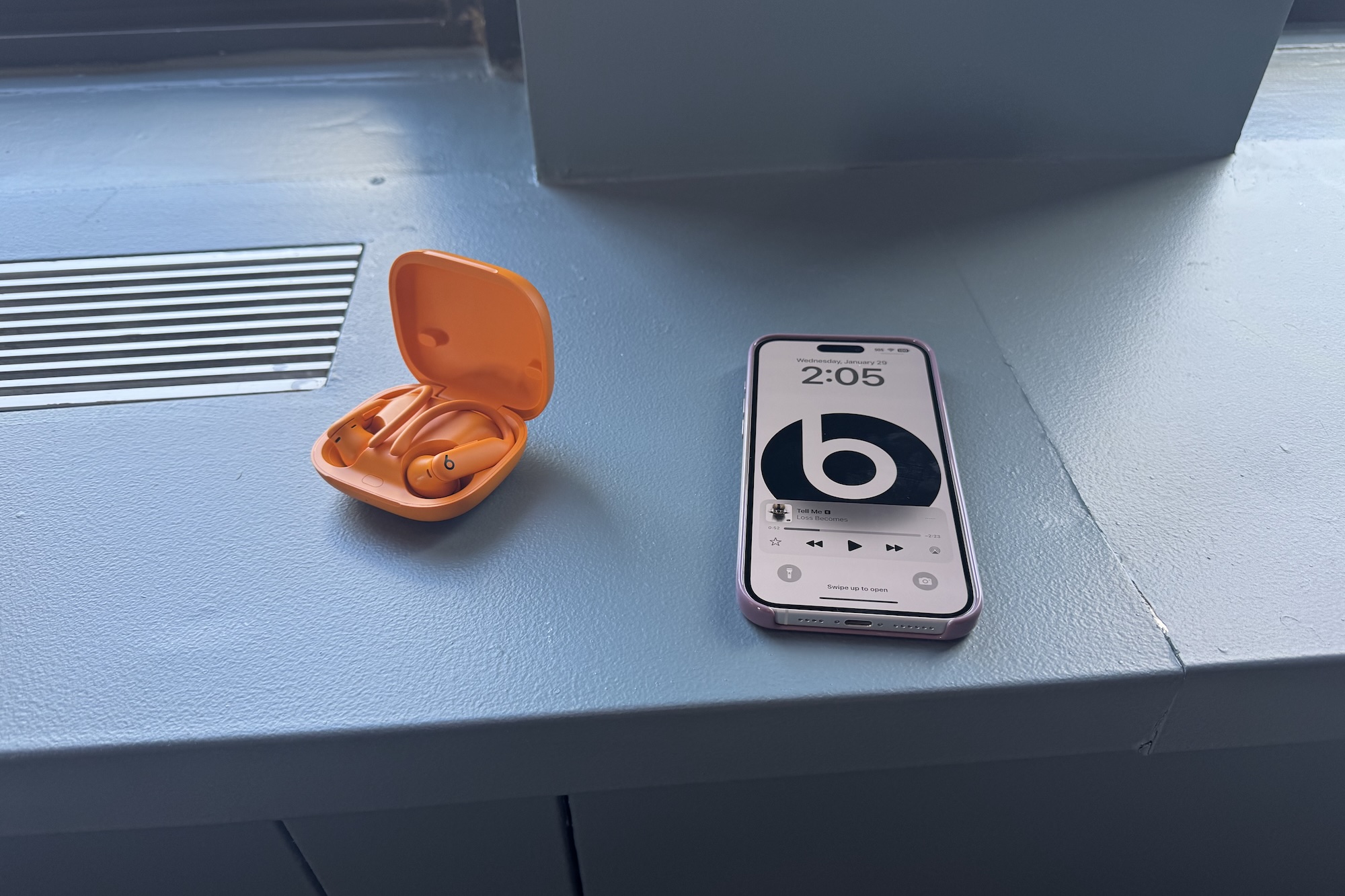 Um par laranja de fones de ouvido Beats Powerbeats Pro 2 em seu estojo ao lado de um iPhone exibindo o logotipo da Beats.