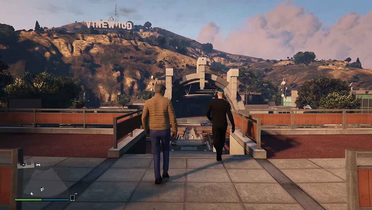 A figure wearing a tan jacket and jeans walks alongside a figure with blue hair wearing all black. They walk down towards a large open ampitheater. A sign that reads "Vinewood" stands behind the venue.