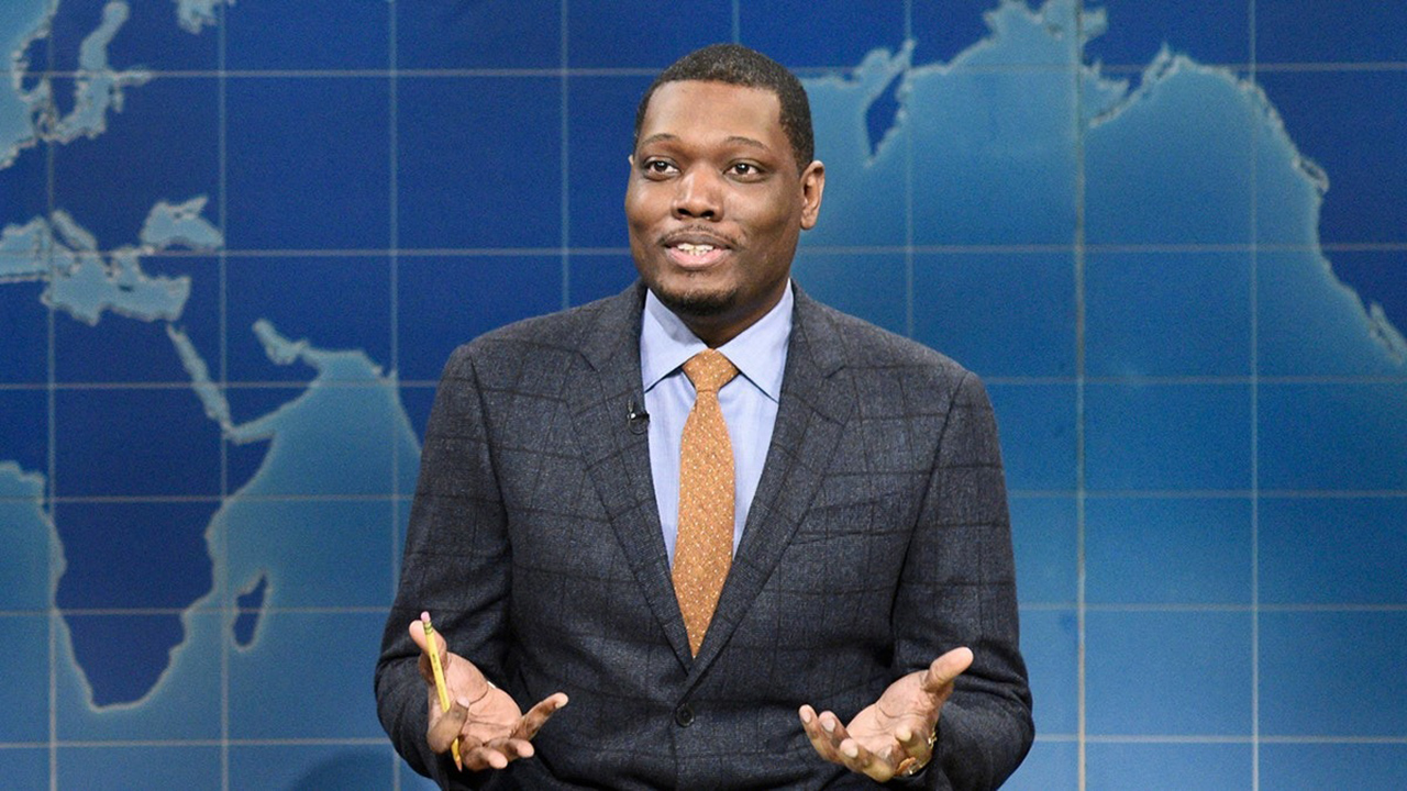 Майкл Че в программе Weekend Update на канале SNL с вытянутыми вперед руками во время разговора.