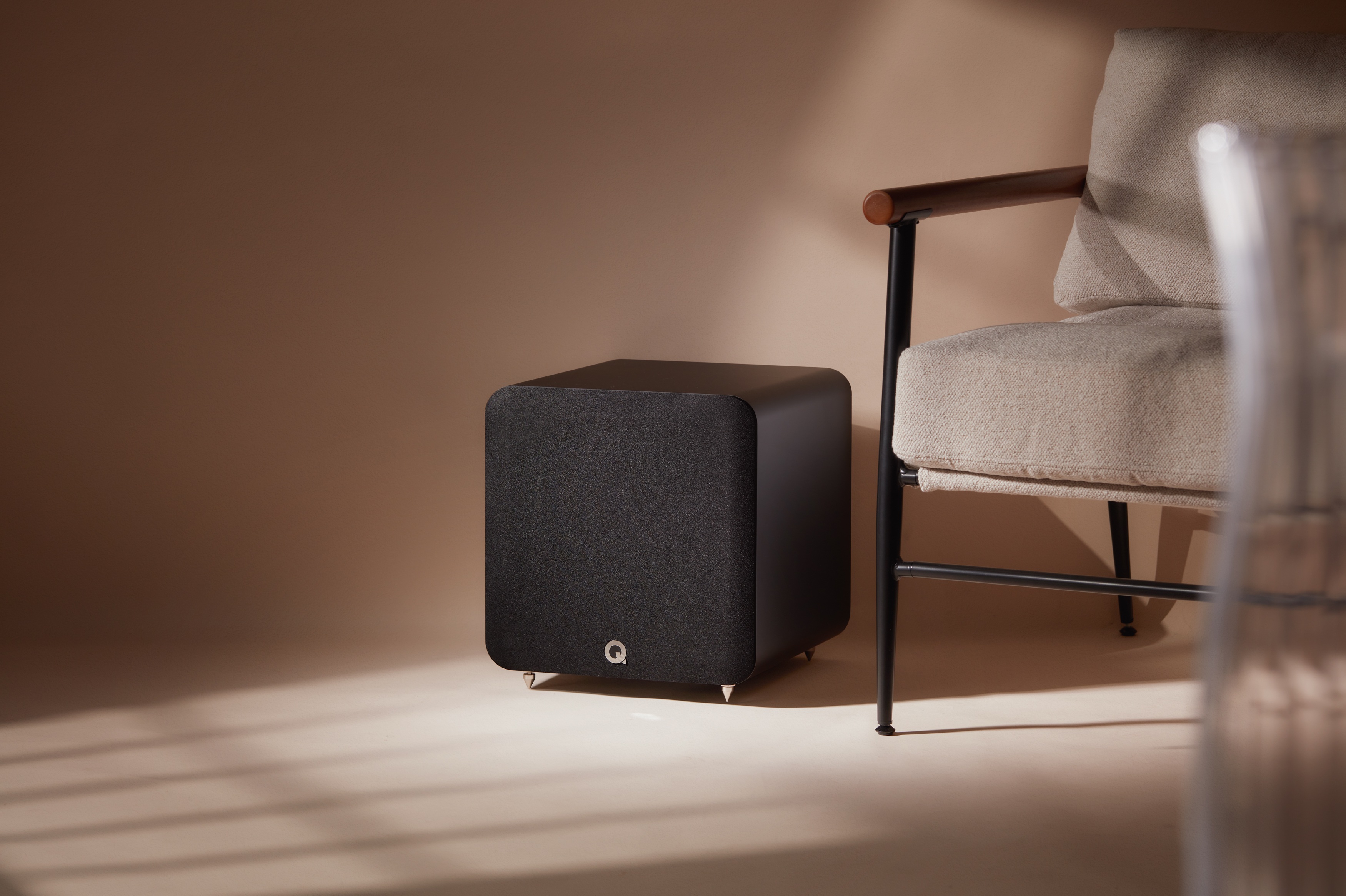 The Q Acoustics SUB100 subwoofer in black, next to a chair.