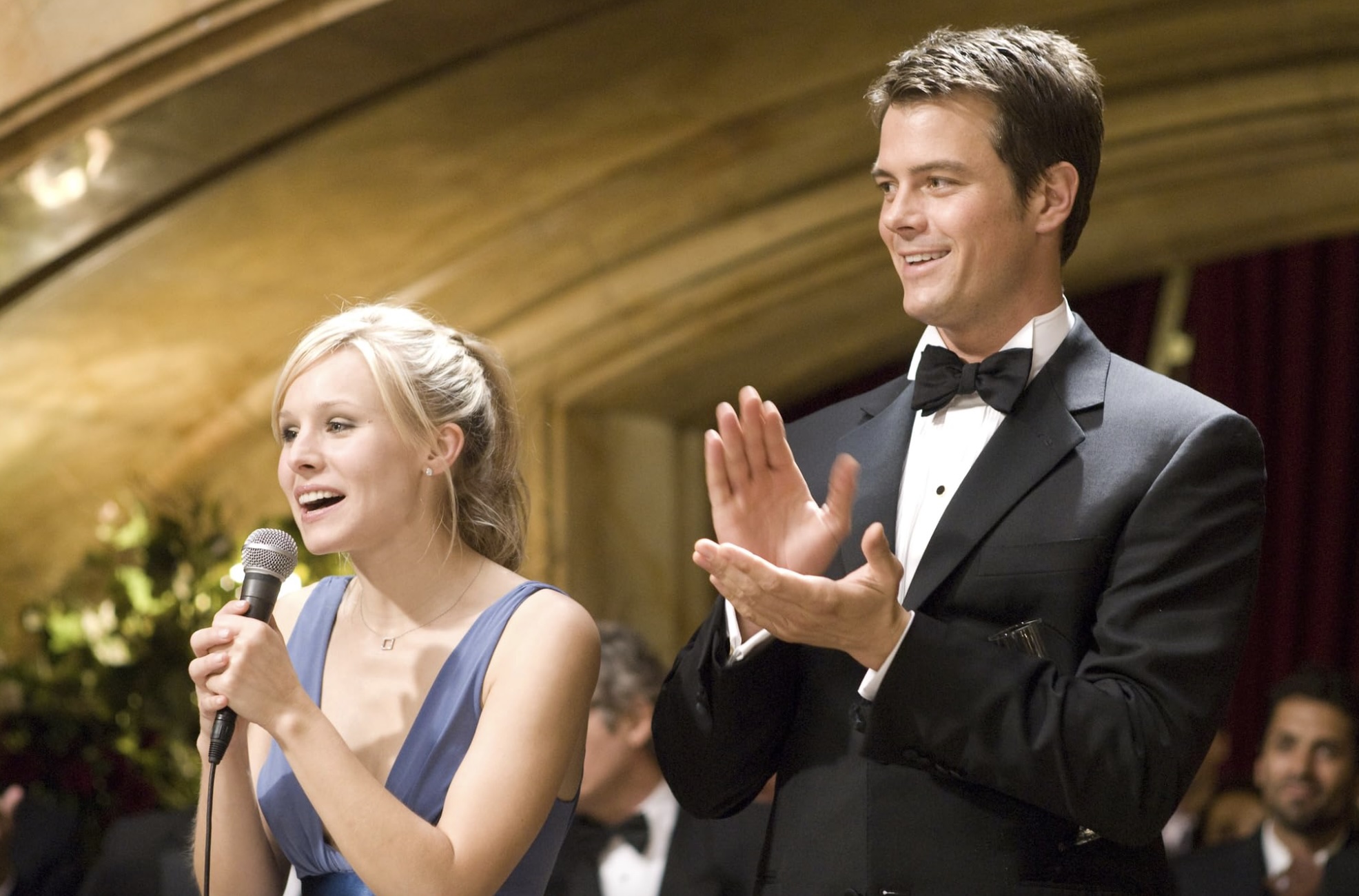 A woman speaks on a mic next to a man clapping.